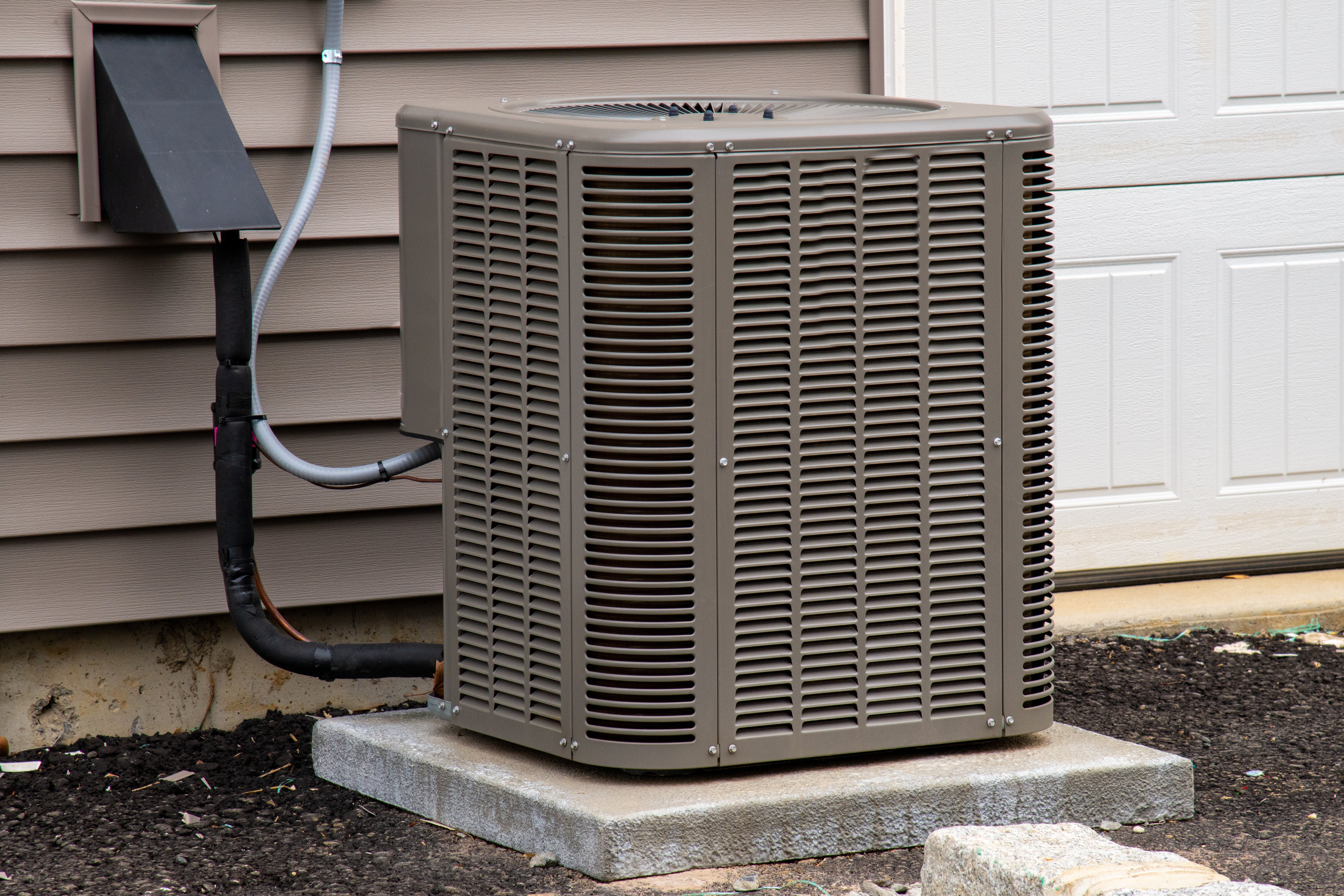 Outdoor air conditioning unit on a concrete pad beside a house, with insulated refrigerant lines and electrical conduit running along the siding.